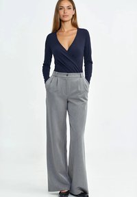 Granatowa bluzka kopertowa z długim rękawem w połączeniu z wysokim stanem jasnoszarych plisowanych spodni. Spodnie mają boczne kieszenie i zapięcie na guzik.