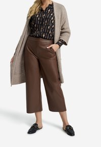 Braune Kunstleder-weite Hose mit Fronttaschen, kombiniert mit einer blauen und beigen gemusterten Bluse und einer hellbeigen Strickjacke.