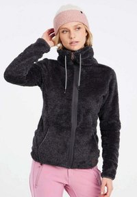 Zwarte fuzzy fleece jas met hoge kraag, ritssluiting aan de voorkant en trekkoordjes. Model draagt een lichtroze gebreide beanie en roze broek.
