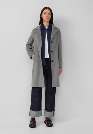 Manteau en laine grise avec un col cranté, deux poches avant et des boutons noirs. Porté par-dessus une chemise en denim bleu marine, un jean foncé et des mocassins marron.