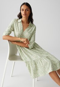 Robe verte à motifs avec un col et des manches trois-quarts, présentant une jupe à niveaux et une texture de tissu douce.