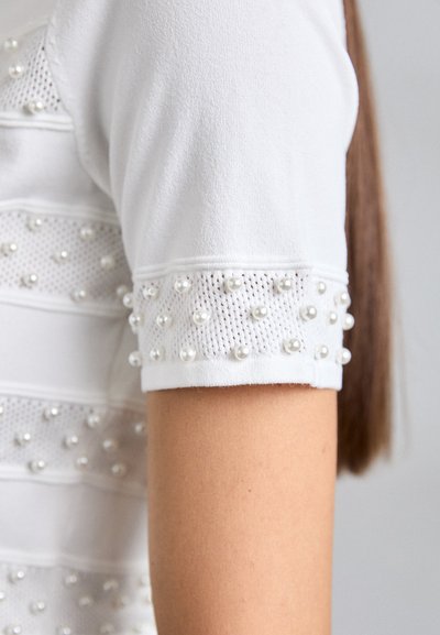 Vêtement à manches courtes blanc avec des perles en forme de perle sur des détails en tissu en maille, porté par une personne ayant de longs cheveux bruns.