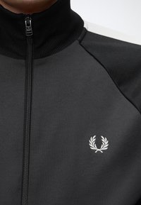 Veste noire zippée avec col côtelé et logo de couronne de laurier blanc sur la poitrine gauche, portée par une personne.