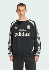 Svart stickad tröja med vitt Adidas-logotyp och ränder. Har rund halsringning och ribbad fåll. Inkluderar en rund emblem på vänster sida.