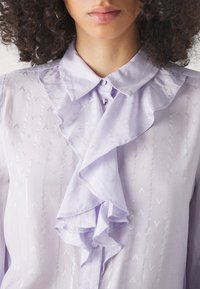 Light lavender blouse with a ruffled collar, button closure, and subtle diagonal patterns embossed on the fabric. Soft texture.