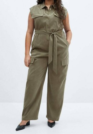 Femme portant une combinaison sans manches vert olive ceinturée avec poches avant et chaussures noires à talons blocs à bout pointu, debout sur un sol blanc.
