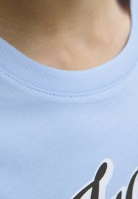 Camiseta de algodón azul claro con cuello redondo ribeteado. Textura suave, presenta letras negras en la parte frontal. Vista en primer plano del escote.