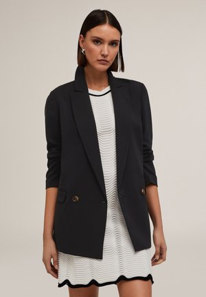 Blazer negro a medida con solapa con muesca, cierre de doble botón y mangas de tres cuartos, llevado sobre un vestido blanco texturizado.