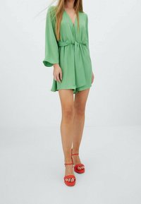 Vestido verde corto con escote en V, cintura fruncida y mangas de campana. Combinado con sandalias de plataforma rojas con correas en el tobillo.