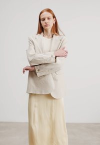 Beige linblazer med hakket revers og avslappet passform, kombinert med en lys gul, flytende maxiskjørt. Enkelt, elegant design med minimalt av maskinvare.