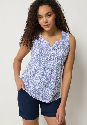 Młoda kobieta o kręconych włosach, ubrana w bezrękawnik w niebiesko-biały wzór oraz granatowe szorty, pozująca z jedną ręką w kieszeni.