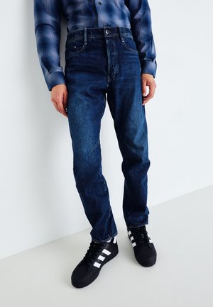 Vaqueros de mezclilla azul con un corte relajado, costuras visibles y una cintura alta; combinados con zapatillas negras y blancas.