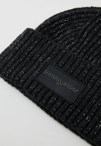 Căciulă tricotată neagră cu fire argintii, având o textură ribrată și un patch din material negru pe care este inscripționat "tommyjeans" în text embossat.