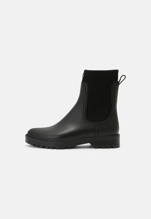 Bota de lluvia negra hasta el tobillo con paneles laterales elásticos acanalados, lengüeta en el talón y suela resistente, mostrada desde el lado exterior sobre un fondo blanco.