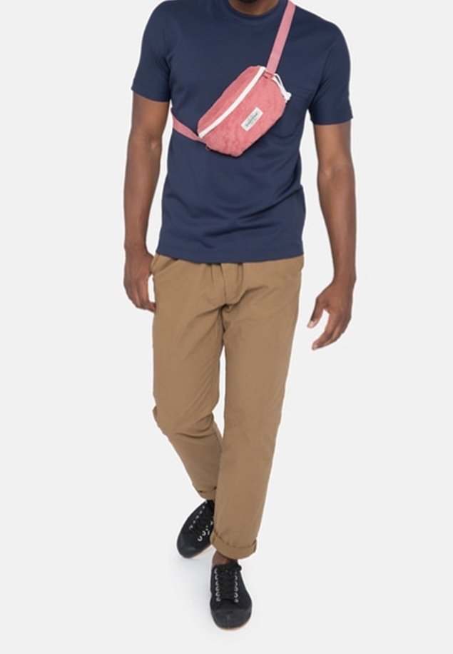 Homme portant un t-shirt bleu marine, un pantalon kaki, des baskets noires et un sac bandoulière rose marchant devant un fond blanc.