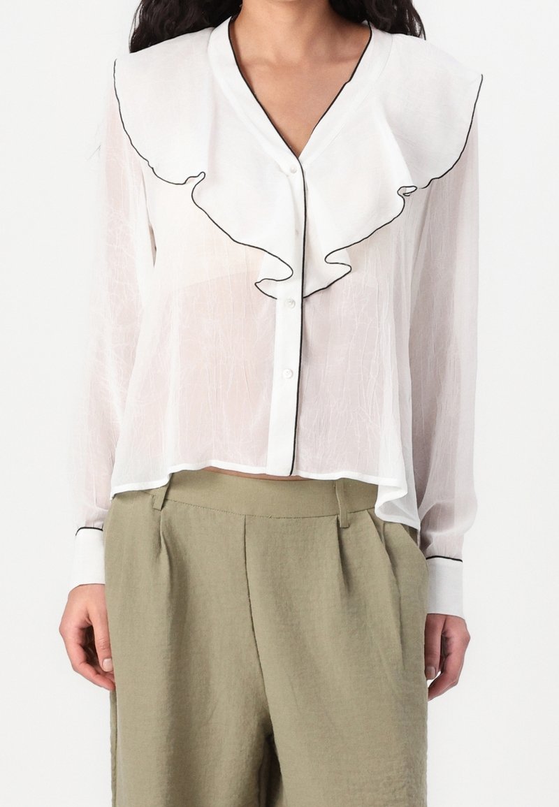 Femme portant un chemisier blanc transparent avec un grand col à volants et une bordure noire, associé à un pantalon beige taille haute.