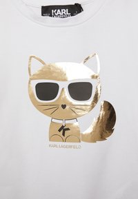 Bílé tričko s ilustrací zlaceného kočky v brýlích. Text "KARL LAGERFELD" vytištěný pod designem.