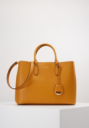 Handtas van tan leer met dubbele handvatten, afneembare schouderriem en een klein decoratief label, geplaatst op een wit voetstuk tegen een effen achtergrond.