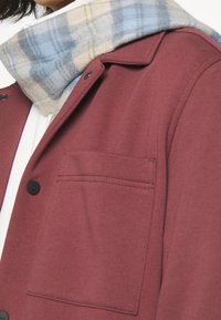 Mörkröd jacka med en texturerad yta, som har en bröstficka och en blå rutig scarf runt kragen. Knappar synliga på framsidan.