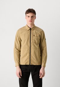 Veste légère beige avec fermeture éclair à l'avant, col et poche poitrine. Présente des coutures renforcées et un ourlet ajustable avec cordon de serrage.