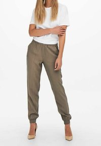 Olijfgroene joggers met een elastische tailleband en boorden, gecombineerd met een wit T-shirt. Model staat in huidskleurige hoge hakken. Eenvoudig ontwerp.