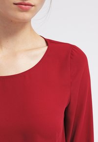 Rote Bluse mit rundem Ausschnitt und langen Ärmeln, aus glattem Stoff mit subtilen Falten an den Schultern.