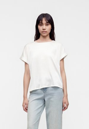 Femme aux cheveux noirs et lisses portant un haut blanc uni à manches courtes et un jean bleu clair, se tenant devant un fond blanc.