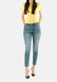 Camisa de manga corta amarilla clara combinada con unos vaqueros de corte slim de tiro medio, de color azul claro, que cuentan con cinco bolsillos y un acabado ligeramente desgastado.