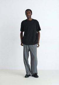 T-shirt en coton noir à manches courtes, associée à un pantalon ample gris en tissu texturé, et des chaussures noires. Fond uni.