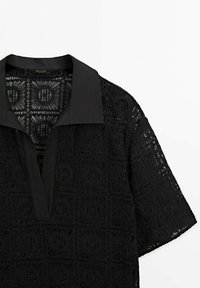 Chemise noire à manches courtes avec un large col et un profond décolleté en V, présentant des motifs complexes en dentelle carrée sur tout le tissu.