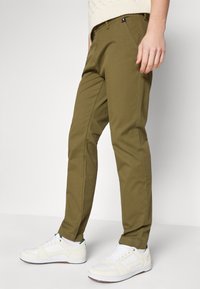 Chino verde oliva con una vestibilità slim, dotati di tasche frontali e una piccola etichetta con logo. Abbinati a sneakers bianche, con una calza in maglia strutturata.