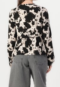 Schwarz-creamfarbige Bluse mit Muster, weitem Saum, langen Ärmeln und rundem Kragen, kombiniert mit grauen hochgeschnittenen Hosen.