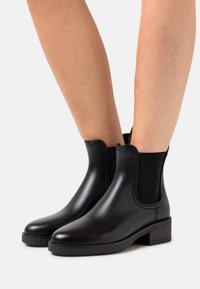 Bottines noires avec une finition lisse et brillante. Panneaux latéraux élastiques et semelle en caoutchouc texturée. Talon carré pour la stabilité et le confort.