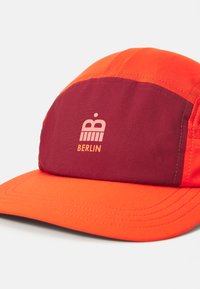 Rød og oransje caps med et bordeaux panel, en grafisk logo, og ordet "BERLIN" i rosa skrift. Glatt stoff med strukturert form.