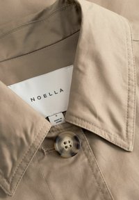 Smėlio spalvos švarkas su apykakle, dideliu vėžlio kiauto mygtuku ir matomu baltu etikete, ant kurios parašyta „NOELLA“ ir „S Made in China“, esančia viduje apykaklės.