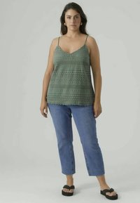 Green lace tank top with thin straps, featuring a textured pattern. Paired with light blue cropped jeans and black sandals.