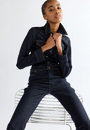 Frau mit geflochtenem Haar, die eine dunkle Jeansjacke und passende Jeans trägt, sitzt auf einem silbernen Metallstuhl vor weißem Hintergrund.