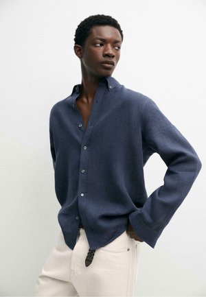 Junger Mann, der ein aufgeknöpftes dunkelblaues Hemd und hellbeige Hose trägt, steht mit einer Hand in der Tasche und blickt zur Seite.