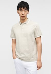 Ανοιχτό μπεζ πόλο πουκάμισο με διακριτικό σχέδιο, κοντά μανίκια και λευκές ρίγες στο γιακά και τις μανσέτες. Φοριέται με λευκό παντελόνι.