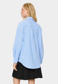 Camicia a maniche lunghe a righe blu chiaro con yoke posteriore e dettagli in occhiello sulle maniche, abbinata a una gonna nera plissettata.