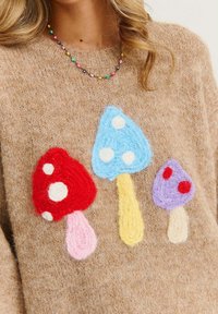 Beżowy sweter z trzema kolorowymi aplikacjami grzybów: czerwonym, jasnoniebieskim i fioletowym, każdy z kropkami i teksturowanymi detalami.