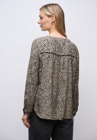 Blusa beige a maniche lunghe con stampa a pois neri, caratterizzata da un dettaglio plissettato sul retro e un collo rotondo. Il tessuto appare morbido e leggero.