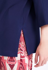 T-shirt blu navy a maniche corte con spacco laterale, abbinato a pantaloni rossi e crema con motivi a spirale e disegni floreali.
