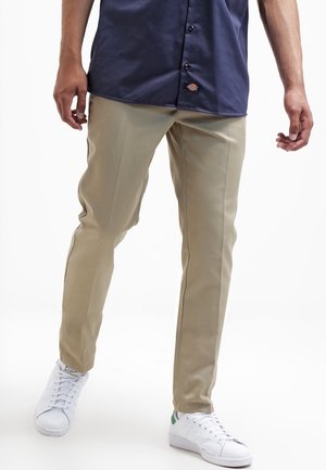 Personne portant un pantalon beige, une chemise bleu marine à boutons et des baskets blanches, debout devant un fond blanc.