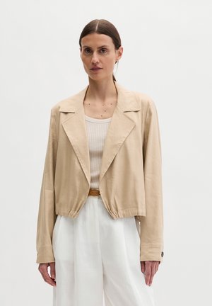 Vrouw met donker haar naar achteren gedragen, draagt een beige cropped jack, een geribde crèmekleurige tanktop, witte broek met hoge taille en een bruine riem.