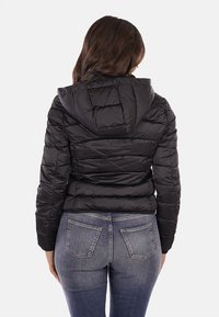 Giacca a vento nera con cappuccio, design trapuntato e finitura testurizzata. La vista posteriore mostra una forma aderente e un orlo elastico, abbinata a jeans blu.