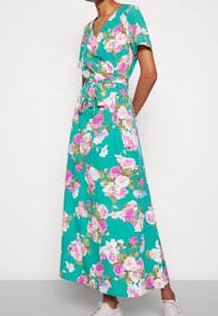 Robe maxi florale en tissu turquoise avec des fleurs roses, blanches et vertes, un décolleté en V, des manches courtes et une ceinture à nouer à la taille.