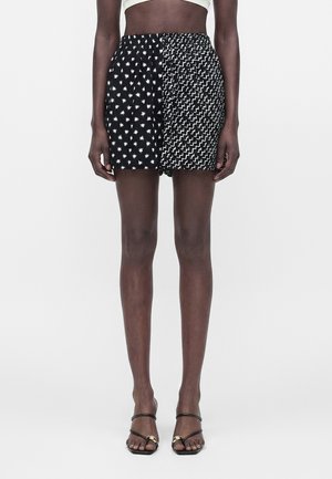 Γυναίκα που φοράει μαύρο σορτς με λευκά αστέρια και ζιγκ-ζαγκ μοτίβα, συνδυασμένο με μαύρα πέδιλα με τακούνια και λουράκια.