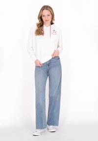 Felpa bianca con coulisse rosa e ricamo floreale sul petto, abbinata a jeans a gamba larga blu chiaro e sneakers bianche.