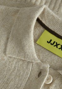 Béžový pletený svetr s měkkou texturou, se skládacím límcem, zapínáním na knoflík a žlutou etiketou s nápisem "JJXX."